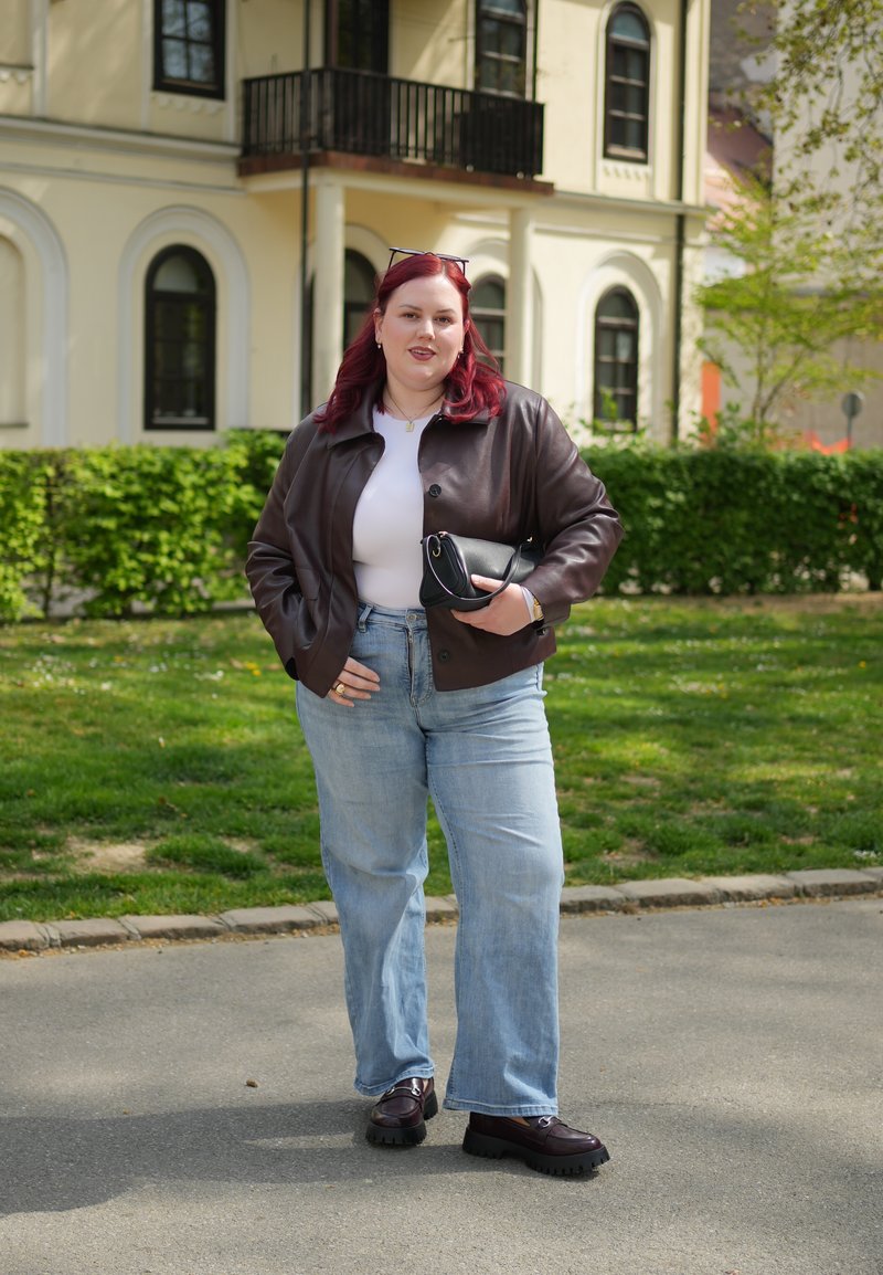 Žena s červenými vlasy má na sobě hnědou koženou bundu, bílý top, modré džíny a černé boty, drží černou kabelku, stojí venku u budovy.