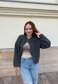 Ung kvinna med brunt hår ler, iklädd en mörkgrå cardigan över en pastellfärgad topp och slitna blå jeans, stående mot en beige stenvägg.