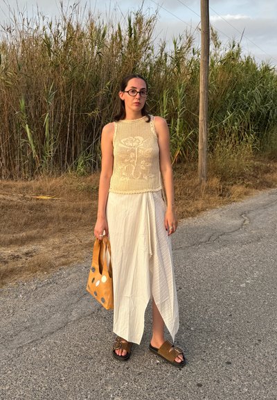 Top de punto beige con un patrón floral, falda blanca plisada, sandalias marrones y un bolso marrón con lunares plateados. De pie en una carretera al lado de hierba alta.