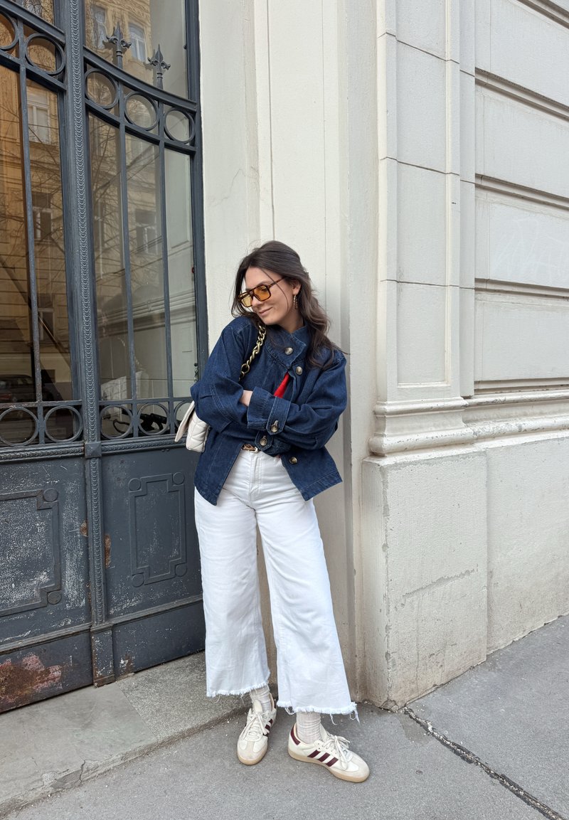 Γυναίκα με γυαλιά ηλίου που φοράει σκούρο μπλε μπουφάν, άσπρο παντελόνι φαρδύ στο κάτω μέρος και αθλητικά παπούτσια, στέκεται με σταυρωμένα τα χέρια στην είσοδο ενός κτιρίου.