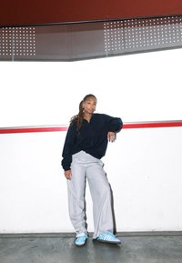 Μαύρο φαρδύ πουκάμισο, ανοιχτό γκρι παντελόνι σε φαρδιά γραμμή και μπλε αθλητικά παπούτσια με λευκές λεπτομέρειες. Το μοντέλο στηρίζεται σε τοίχο με κόκκινες λεπτομέρειες.