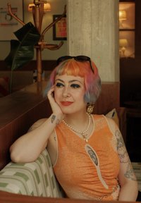 Oranje mouwloze jurk met gerimpelde details, parelketting en grote oorbellen. Achtergrond toont een groene plant en binnenverlichting.