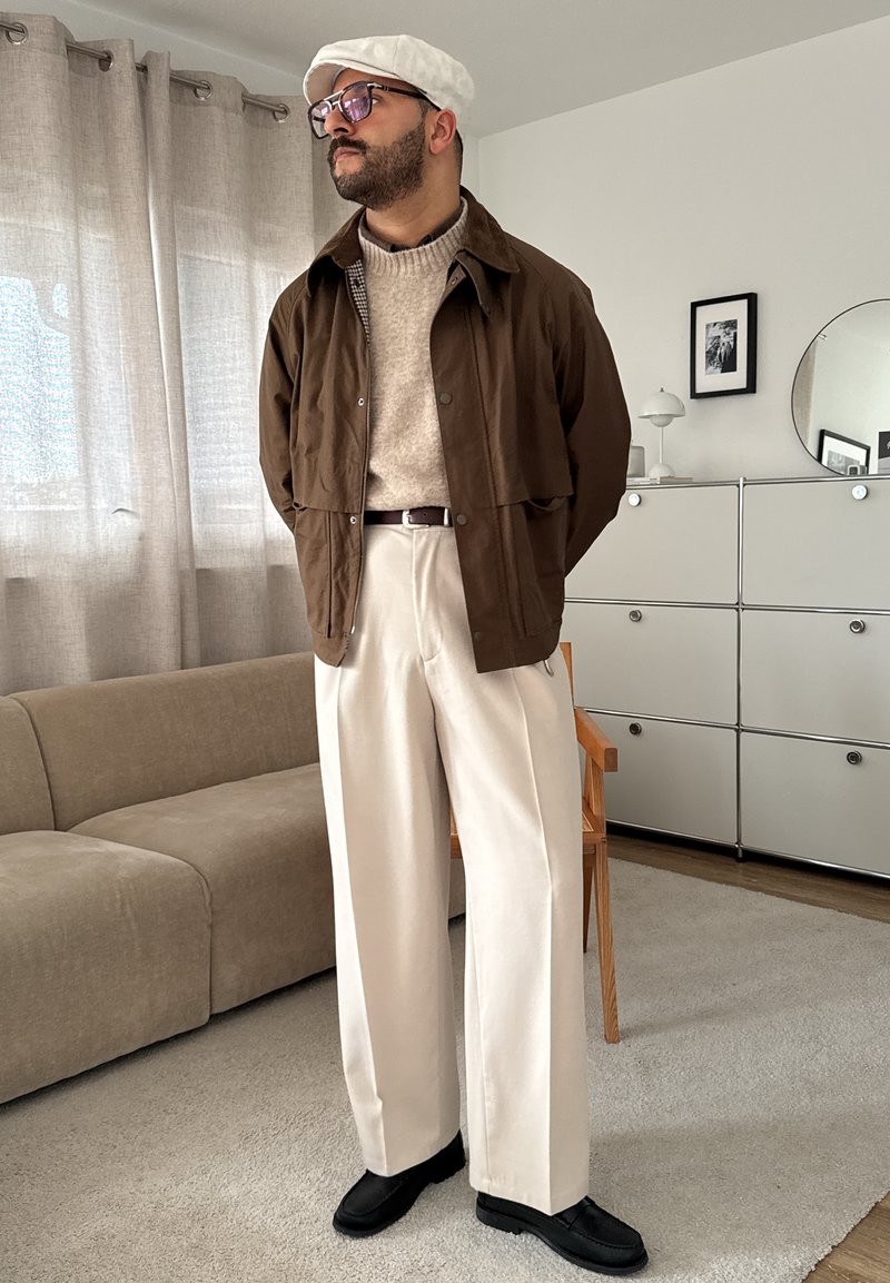 Brun overshirt, beige striktrøje, brede cremefarvede bukser, sorte sko og en hvid kasket. Rummet har bløde teksturer og neutrale farver.