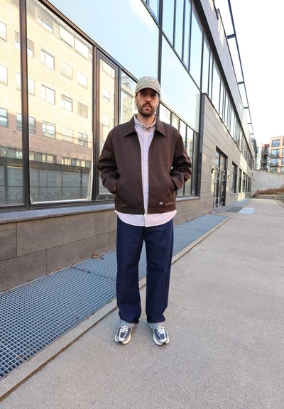 Hombre con chaqueta marrón, camisa clara, vaqueros azules, zapatillas grises y gorra beige, de pie en la acera junto a un edificio de cristal.