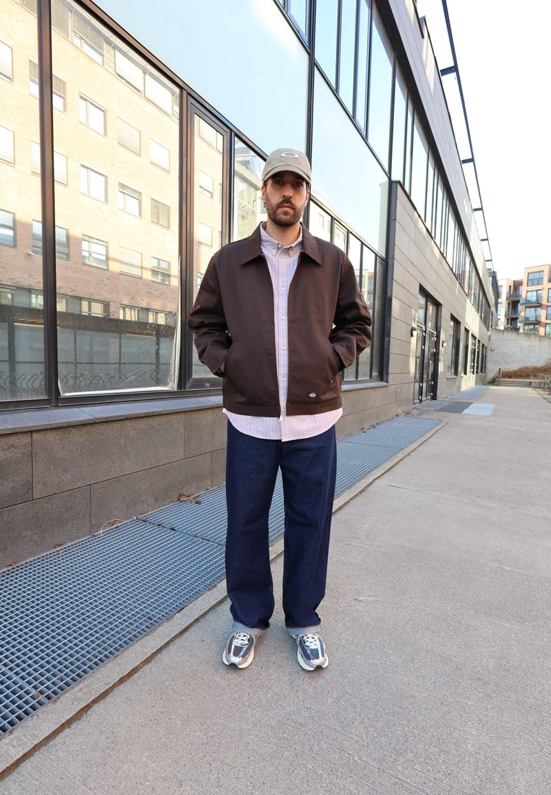 Bărbat purtând o jachetă maro, cămașă deschisă, blugi albaștri, adidași gri și șapcă bej, stând pe trotuar lângă o clădire de sticlă.