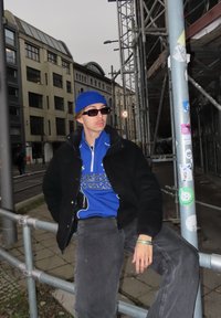 Blå lue, mørke solbriller, svart jakke over en blå zip-up racinginspirert genser, grå bukser; sitter på et metallrekkverk i urbant miljø.