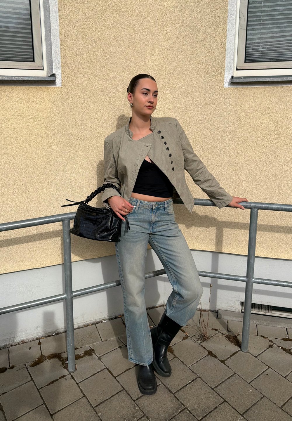 Mujer apoyada en una barandilla de metal afuera, vestida con chaqueta beige, top corto negro, vaqueros azul claro, botas negras y sujetando un bolso negro.