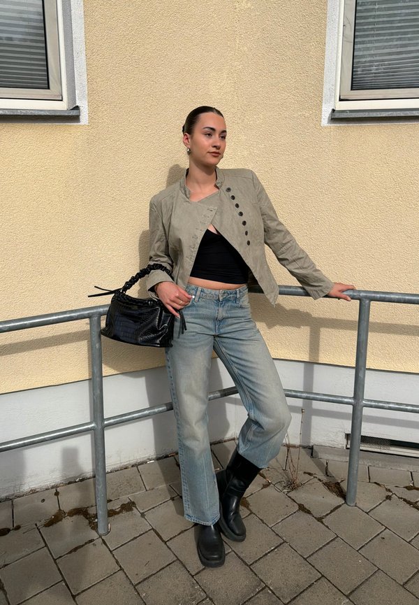 Mujer apoyada en una barandilla de metal afuera, vestida con chaqueta beige, top corto negro, vaqueros azul claro, botas negras y sujetando un bolso negro.