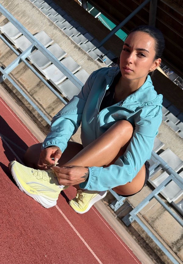 Athlete on a track wearing a light blue zip-up jacket and yellow sports shoes, tying the laces. The background features empty stands.