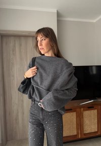 Kvinne med langt brunt hår iført stor grå genser og svarte jeans med nagler, som holder en svart håndveske, står innendørs nær et trebord.