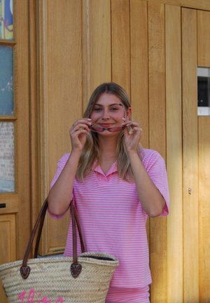Una mujer que lleva una camiseta polo de rayas rosas sostiene unas gafas de sol, con una bolsa tejida a su lado. El fondo presenta paneles de madera y una ventana.
