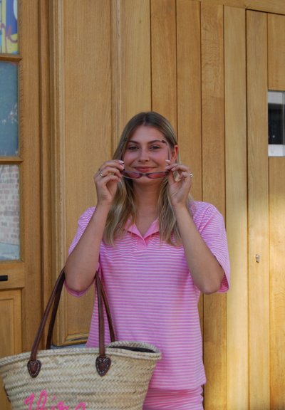 Una mujer que lleva una camiseta polo de rayas rosas sostiene unas gafas de sol, con una bolsa tejida a su lado. El fondo presenta paneles de madera y una ventana.