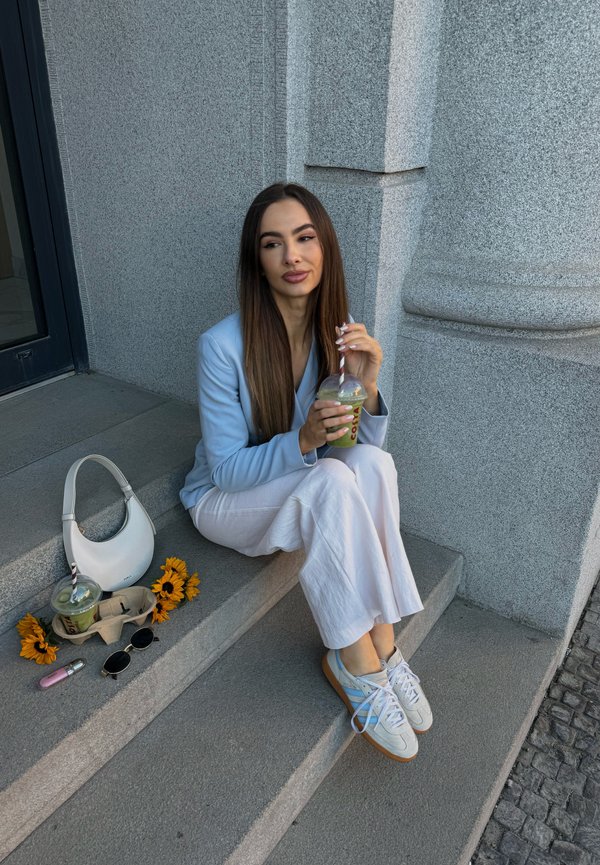 Jeune femme en blazer bleu clair et pantalon blanc assise sur des marches en pierre, buvant une boisson froide, avec un sac à main blanc, des lunettes de soleil et des tournesols à proximité.