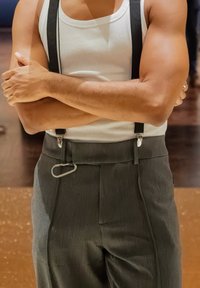 Calças cinzentas texturizadas com cintura alta, apresentando suspensórios pretos e um detalhe de gancho metálico. Usadas com uma t-shirt branca.