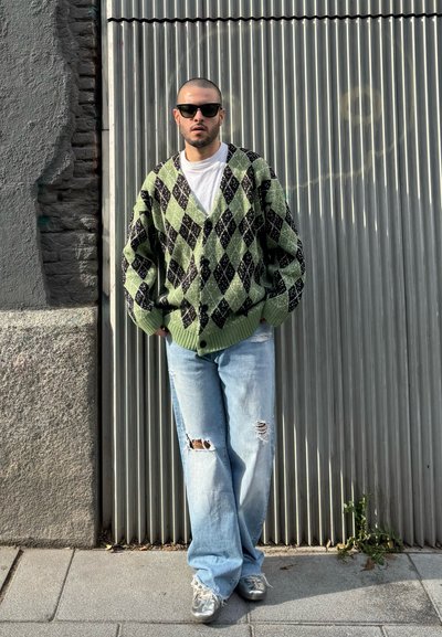 Cárdigan oversize de patrón argyle en verde y negro, camiseta blanca, jeans claros rasgados y zapatillas plateadas. Lleva puestas unas gafas de sol negras.