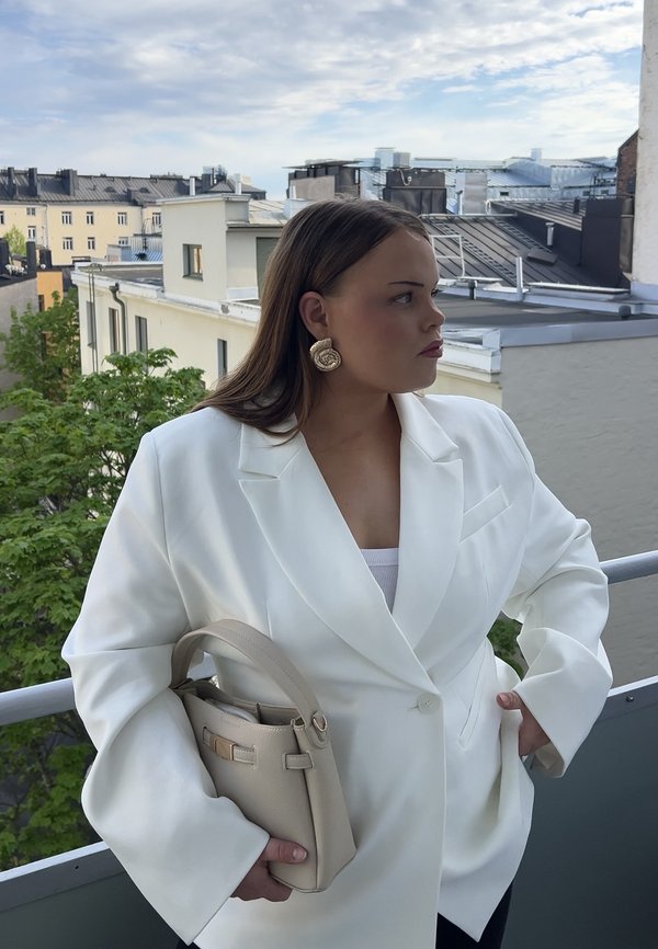 Frau im weißen Blazer hält eine beigefarbene Handtasche und steht auf einem Balkon mit Blick auf städtische Dächer unter einem bewölkten Himmel.
