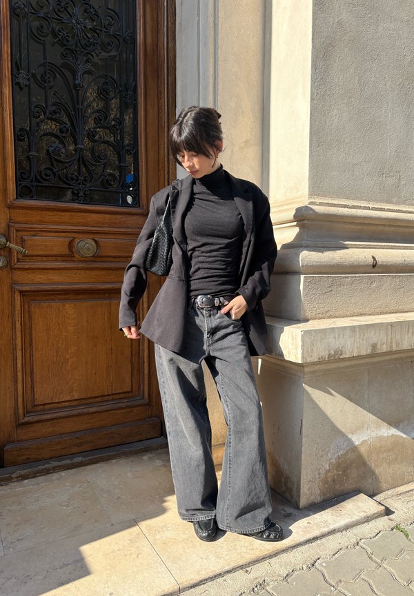 Femme portant un col roulé noir, un blazer, un jean gris à jambes larges et des chaussures noires, debout près d'une porte en bois et d'un mur en pierre au soleil.