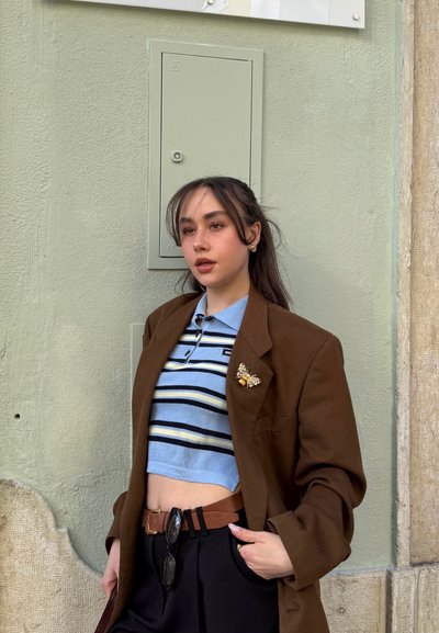 Blazer marrón sobre un polo corto a rayas azules y blancas, cinturón marrón, pantalones negros y un broche de mariposa. Fondo de pared en un verde suave.