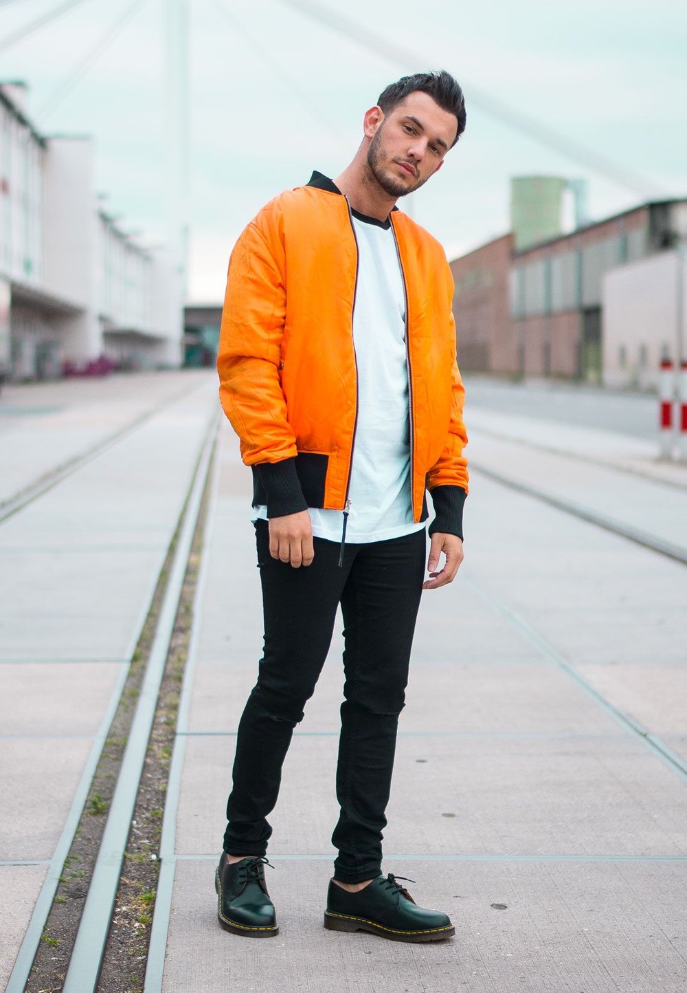 Orangene Bomberjacke mit schwarzen Akzenten  über einem weißen T-Shirt, kombiniert mit engen schwarzen Hosen und schwarzen Schnürschuhen. Urbanes Umfeld.
