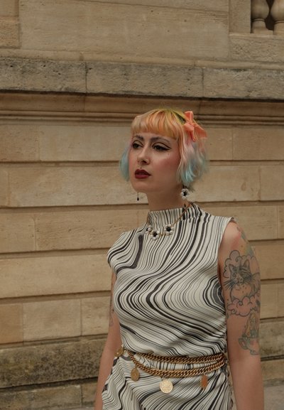 Vestido sin mangas con un patrón ondulado en blanco y negro, cuello alto y cinturón de cadena dorada. Los accesorios incluyen un collar y pendientes de perla.