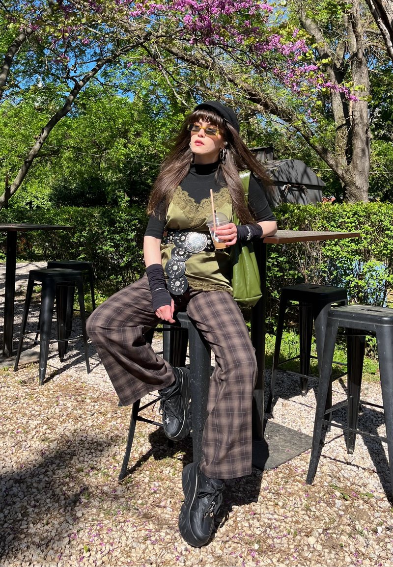 Personne portant un pantalon marron à motifs, une chemise noire, un débardeur vert, un béret noir et des lunettes de soleil assise sur un tabouret extérieur tenant une boisson glacée sous un arbre en fleurs.