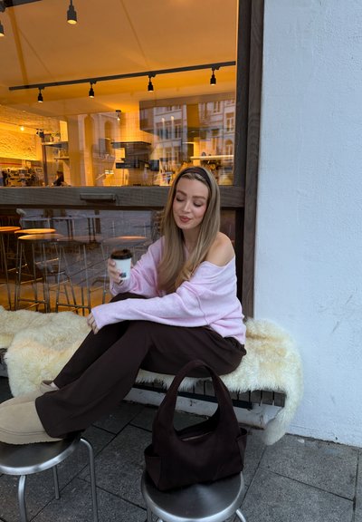 Mujer con suéter rosa y pantalones marrones sentada en un banco cubierto con piel, sosteniendo una taza de café afuera de una cafetería, bolso negro en un taburete cercano.