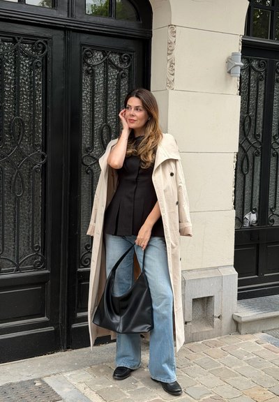 Mujer con gabardina beige y top negro está apoyada contra una puerta decorativa de hierro negro, sosteniendo un gran bolso negro en una calle empedrada.