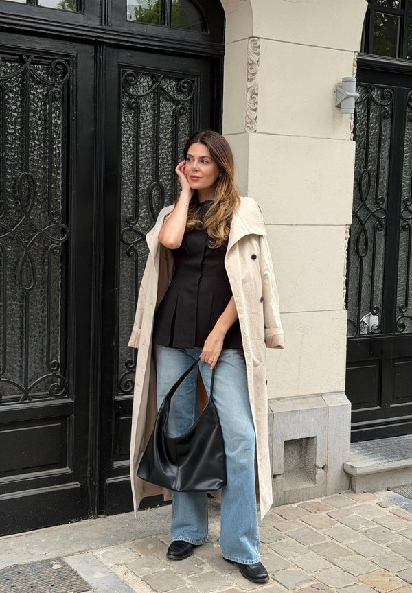 Femme en trench beige et haut noir debout contre une porte en fer noire décorative, tenant un grand sac à main noir dans une rue pavée.