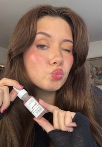 Przezroczysta butelka z zakraplaczem i białą nakrętką, zawierająca jasno różowe serum. Użytkownik mruga okiem, trzymając butelkę w jednej ręce, na neutralnym tle.