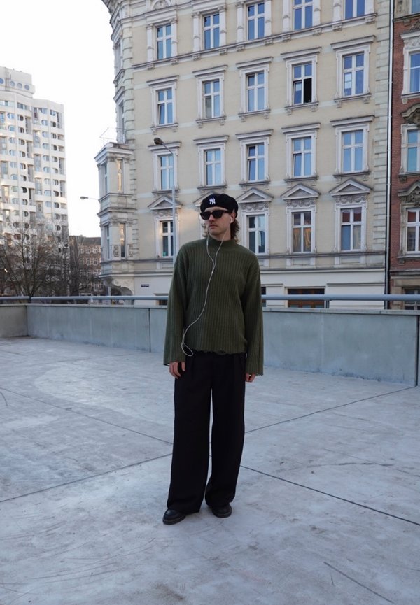 Personne portant un béret noir et des lunettes de soleil, un pull vert, un pantalon large noir, debout sur une terrasse en béton avec des bâtiments urbains en arrière-plan.