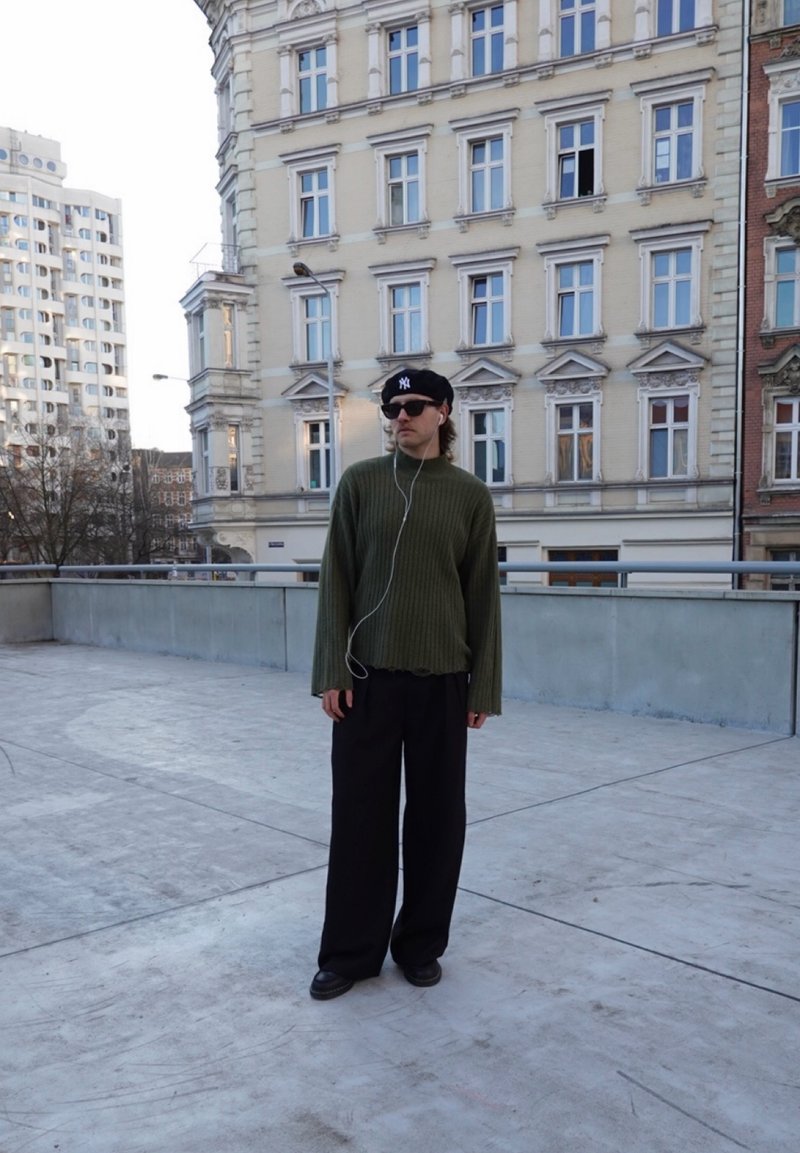 Persoon met een zwarte baret en zonnebril, groene trui, zwarte wijde broek, staande op een betonnen terras met stedelijke gebouwen op de achtergrond.