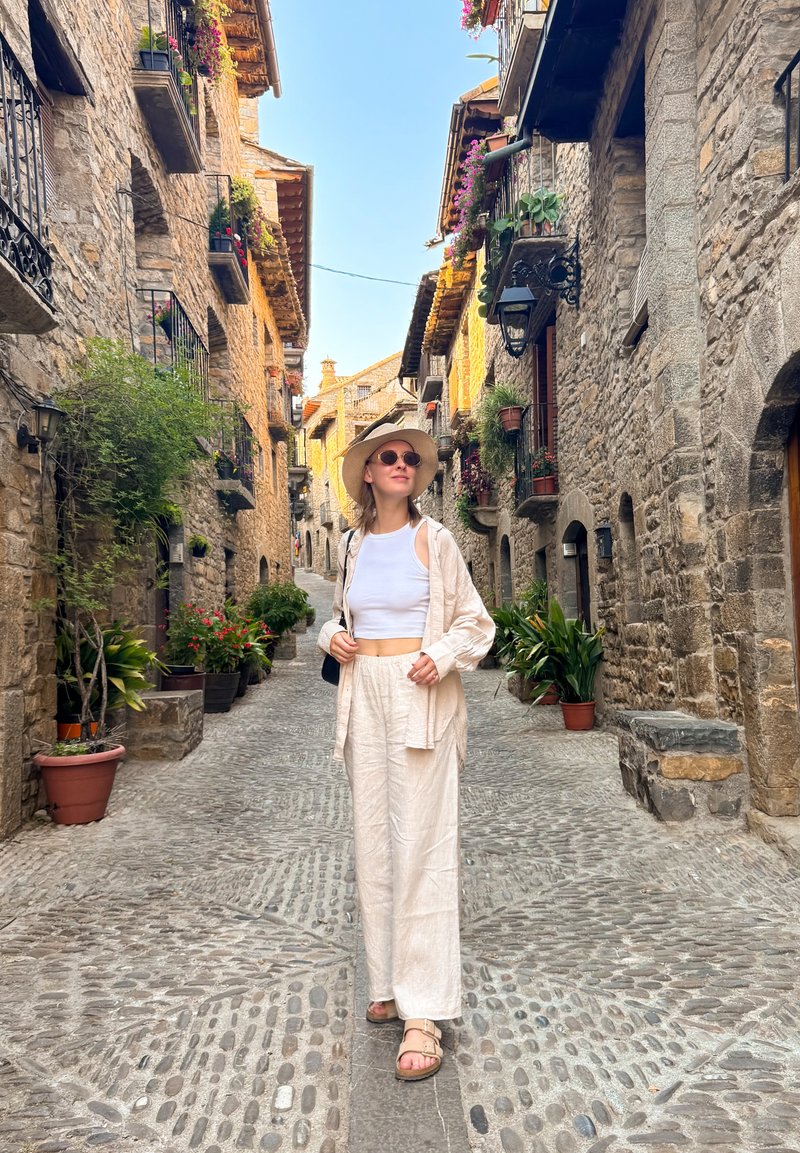 Lightweight, cream-colored linen outfit with a cropped top and loose pants. Wide-brimmed hat, round sunglasses, and a black bag. Stone street alley.
