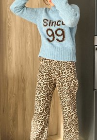 Γαλάζιο υφασμάτινο πουλόβερ με την ένδειξη "Από το 99" σε καφέ, συνδυασμένο με μπεζ παντελόνι με λεοπάρ σχέδιο. Φόντο ξύλου και σύγχρονη συσκευή ορατή.