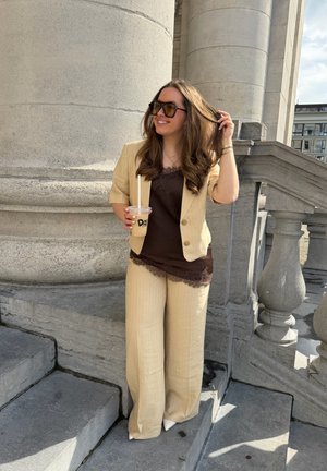 Joven mujer con traje beige y top oscuro está de pie sobre escalones de piedra, sosteniendo un café con hielo y ajustándose el cabello cerca de grandes columnas del edificio.