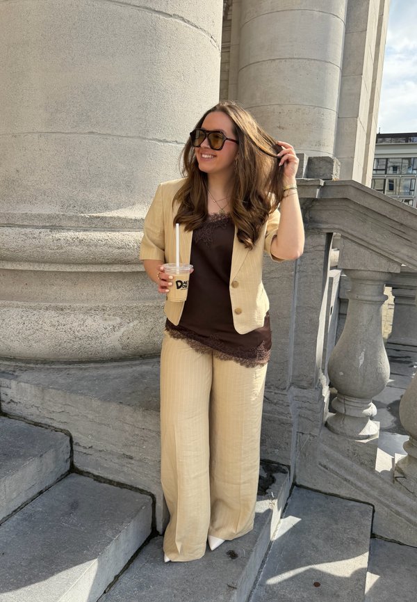 Jeune femme en costume beige et haut sombre se tient sur des marches en pierre, tenant un café glacé et ajustant ses cheveux près de grandes colonnes de bâtiment.