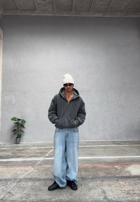 Šedá mikina na zip s kapucí, světle modré kalhoty s širokými nohavicemi, černé boty a bílá čepice, vše na pozadí jednoduché šedé zdi.