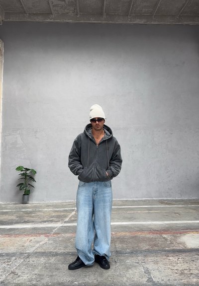 Sudadera gris con cremallera y capucha, vaqueros anchos de pierna en azul claro, zapatos negros y un gorro blanco, sobre un fondo de pared gris lisa.