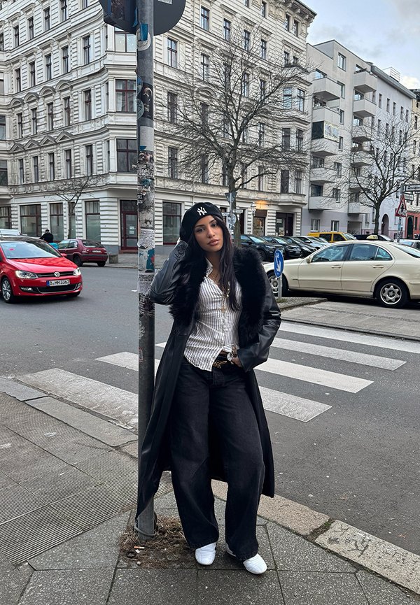 Cappotto nero con bordo in pelliccia sintetica sopra una camicia bianca a righe, jeans larghi neri e sneakers bianche. La scena include segnali stradali e auto parcheggiate.