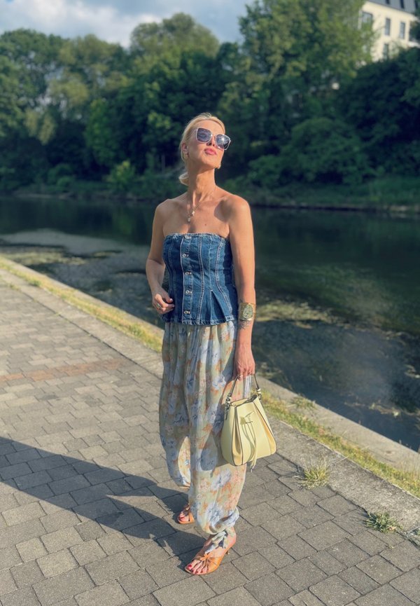 Denim top brez naramnic s gumbi, lahkotna pastelna cvetna krila, prosočna sončna očala, rumena torbica in bež japonke na kamniti poti.