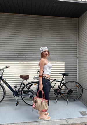 Bolso de mano tejido en color beige con acentos marrones, superfície texturada, llevando una pequeña muñeca. Modelo con una camiseta sin mangas blanca, jeans oscuros y chanclas.