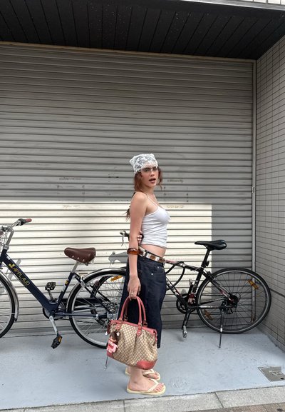 Bolso de mano tejido en color beige con acentos marrones, superfície texturada, llevando una pequeña muñeca. Modelo con una camiseta sin mangas blanca, jeans oscuros y chanclas.