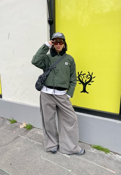 Mujer ajustándose las gafas de sol, con chaqueta verde, pantalones anchos grises, gorra y bolso negro cruzado, de pie junto a una pared amarilla con arte de árbol en forma de corazón.