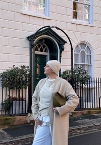 Personne vêtue d'un manteau beige et d'un bonnet en tricot tenant une pochette marron, debout avec assurance sur une rue pavée devant une porte verte et un bâtiment blanc.