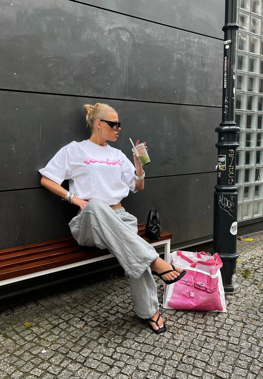 Λευκή κοντομάνικη μπλούζα με ροζ κείμενο, γκρίζες φόρμες, μαύρες σαγιονάρες και μαύρη τσάντα. Πράσινο ποτό σε διάφανο ποτήρι. Ροζ και λευκή τσάντα κοντά.