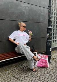 Λευκή κοντομάνικη μπλούζα με ροζ κείμενο, γκρίζες φόρμες, μαύρες σαγιονάρες και μαύρη τσάντα. Πράσινο ποτό σε διάφανο ποτήρι. Ροζ και λευκή τσάντα κοντά.