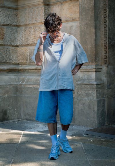 Camisa corta de manga corta color beige claro, pantalones cortos de mezclilla azules, calcetines blancos y zapatillas azul claro. Pared texturizada en el fondo.