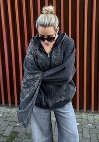 Sweat à capuche noir oversize avec fermeture éclair et accents en strass, doté d'une capuche et de manches larges, associé à un pantalon large gris clair.