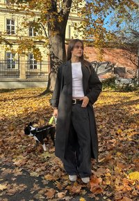 Abrigo negro estilo trench sobre camiseta blanca, pantalones anchos de mezclilla oscuros, zapatos grises. De pie sobre hojas caídas con un perro blanco y negro con correa.