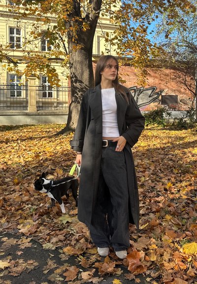 Abrigo negro estilo trench sobre camiseta blanca, pantalones anchos de mezclilla oscuros, zapatos grises. De pie sobre hojas caídas con un perro blanco y negro con correa.