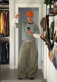 Gestreept T-shirt met korte mouwen in groen en wit, gecombineerd met loszittende olijfgroene broek. Oranje pet, staand voor een spiegel.